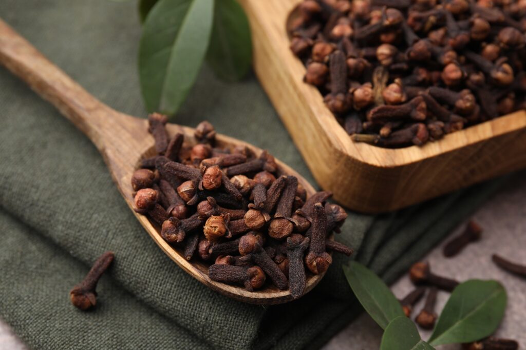 Bowl with aromatic cloves, spoon and green leaves on table. Image Via Shutterstock