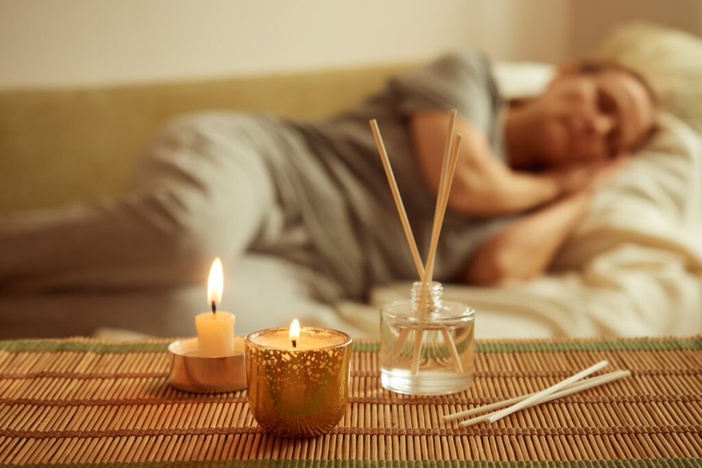 A scented candle, diffuser, and a sleeping woman in the background. Image Via Shutterstock