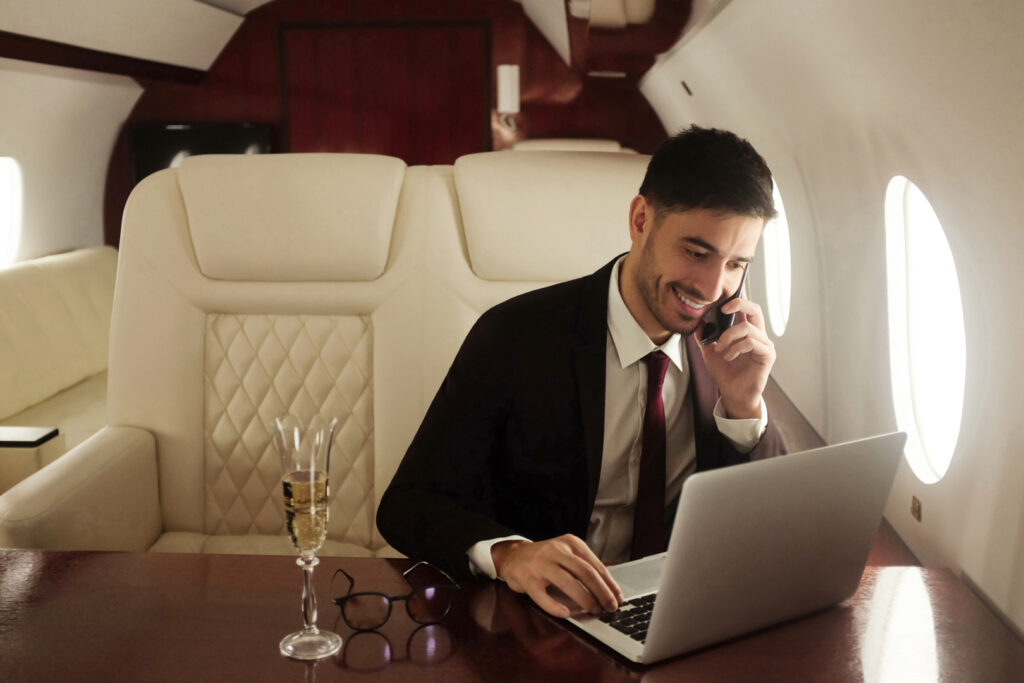 Multimillonario o rico hombre de negocios volando en primera clase y trabajando en el avión con portátil y copa de champán. Imagen Via Shutterstock